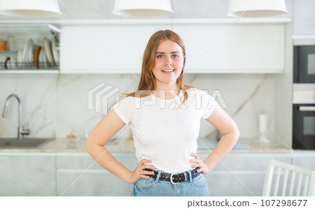 Portrait of long-haired young woman inwhite T-shirt, standing in kitchen Portrait of long-haired young woman inwhite T-shirt, standing in kitchen 107298677