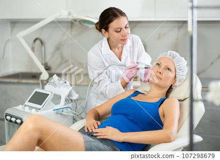 Professional female cosmetologist doing hydrafacial procedure in cosmetology clinic. Doctor use hydrogen and oxygen small bubbles for deeply clear skin. Rejuvenation and hydratation 107298679