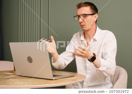 Puzzled business man sit at table, looking at laptop screen with misunderstood eyes. Dazed human raised hands in shock. 107301972