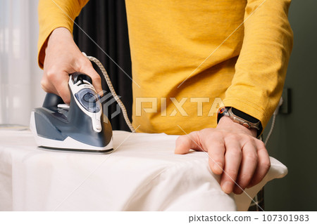 Close up of adult man with watch on hand iron white shirt on pressboard using electric flatiron. Male doing housework. Close up of adult man with watch on hand iron white shirt on pressboard using electric flatiron. Male doing housework. 107301983