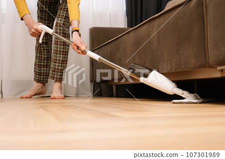Unrecognizable adult human barefoot wearing plaid pants and yellow longsleeve washing floor under couch in living room. Unrecognizable adult human barefoot wearing plaid pants and yellow longsleeve washing floor under couch in living room. 107301989