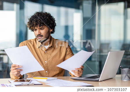 Serious and upset businessman checking and analyzing documents financial reports, thinking hispanic man reviewing contracts on paper work, man inside office at workplace with laptop. 107303107