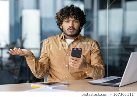 Portrait of unhappy and sad businessman at workplace inside office, man looking at camera holding phone in hands, using app, received spam message and blocked smartphone. Portrait of unhappy and sad businessman at workplace inside office, man looking at camera holding phone in hands, using app, received spam message and blocked smartphone. 107303111