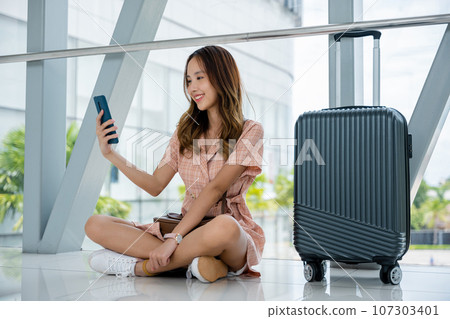 Happy woman smiling and texting on her smartphone while waiting for her flight at the airport. Tourist journey trip concept 107303401