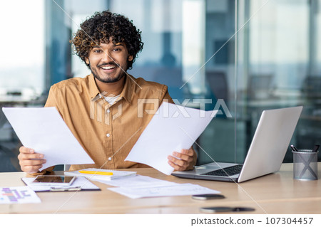 Portrait of young successful financier businessman paper work, hispanic smiling and looking at camera man holding paper reports and bills in hands, satisfied with achievement and checking results. 107304457