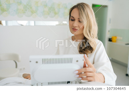 Portrait of professional female beautician typing on screen of device in beauty salon during aesthetic procedure. Concept of non-surgery cosmetology, professional skincare, beauty treatment Portrait of professional female beautician typing on screen of device in beauty salon during aesthetic procedure. Concept of non-surgery cosmetology, professional skincare, beauty treatment 107304645