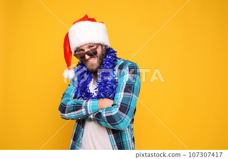 bearded man in shirt, santa hat, boa and sunglasses smiling on yellow background 107307417