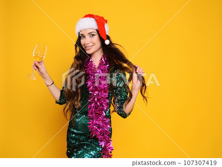 pretty woman in green sparkly cocktail dress wearing santa hat holding pink boa and champagne glass smiling on yellow background 107307419