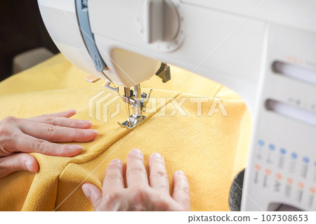 Tailor hands stitching yellow fabric on modern sewing machine at workplace in atelier Tailor hands stitching yellow fabric on modern sewing machine at workplace in atelier 107308653