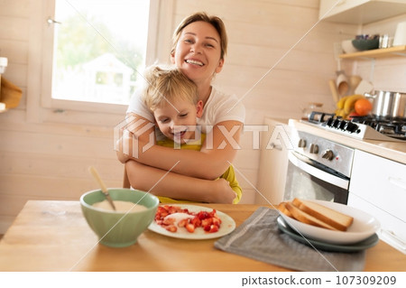 cheerful Caucasian blond mother and child son are sitting in the kitchen and eating. happy childhood cheerful Caucasian blond mother and child son are sitting in the kitchen and eating. happy childhood 107309209