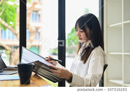 Asian female financier reading paper document, analyzing marketing data at desk in bright office 107310283