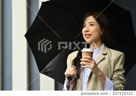 Elegant millennial businesswoman with umbrella and disposable paper coffee cup walking down city street 107310322