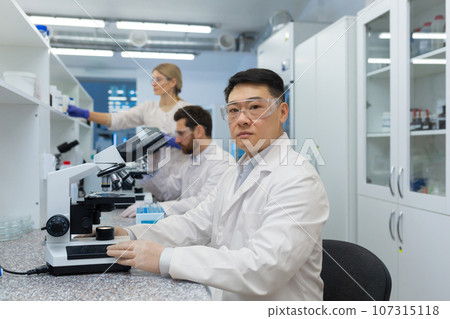 A group of young medical students and scientists are studying in the laboratory of the university. Asian young man sits behind a microscope and looks seriously at the camera. A group of young medical students and scientists are studying in the laboratory of the university. Asian young man sits behind a microscope and looks seriously at the camera. 107315118
