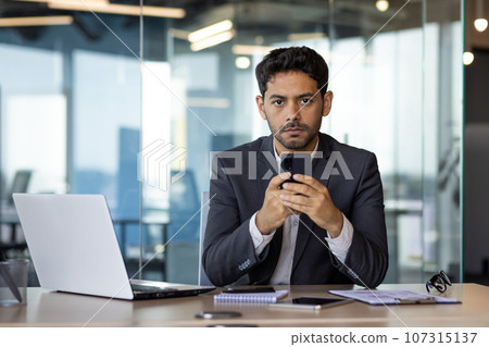Portrait of a young serious man businessman, executive director sitting in the office at the desk and using the phone, confidently looking at the camera. 107315137