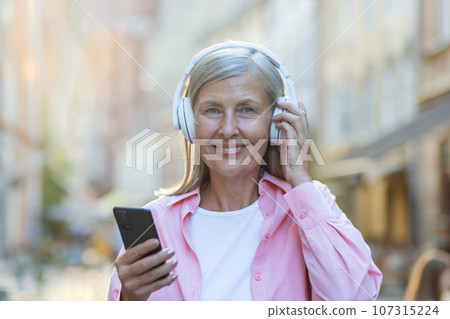 Portrait of an older beautiful gray-haired woman wearing headphones listening to music on the phone. Standing on a city street and smiling at the camera. Close-up photo. Portrait of an older beautiful gray-haired woman wearing headphones listening to music on the phone. Standing on a city street and smiling at the camera. Close-up photo. 107315224