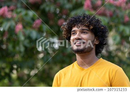 Close-up photo. Portrait of a young Indian man standing in an orange t-shirt outside in a park and looking to the side smiling. 107315233