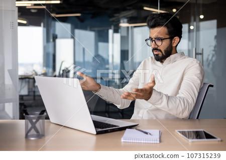 Upset young Indian businessman man working in office and looking frustrated at laptop while spreading hands. 107315239