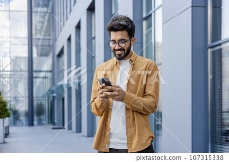 A young smiling man walks through the city with a phone in his hands, outside an office building, happily uses an application on a smartphone, reads messages, types, and browses the Internet. 107315338
