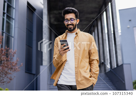 A young smiling man walks through the city with a phone in his hands, outside an office building, happily uses an application on a smartphone, reads messages, types, and browses the Internet. 107315340
