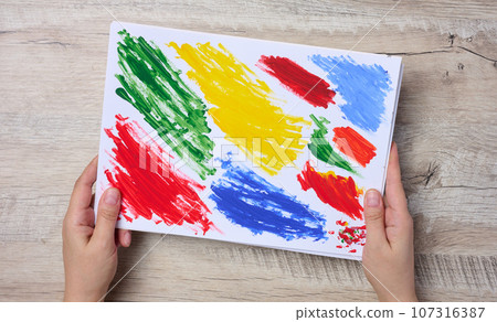 Female hands holding a white sheet of paper with strokes of acrylic multi-colored paint, top view 107316387