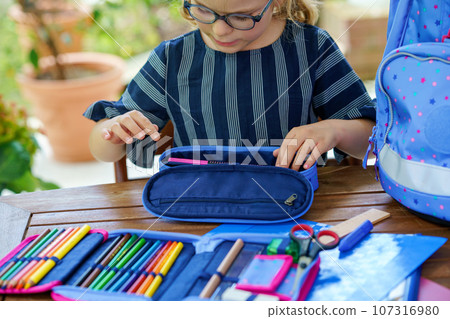 Happy smiling girl preparing for school her backpack. First day of school. Back to school concept. Little child collecting different supplies like pens and books in a bag or satchel. 107316980