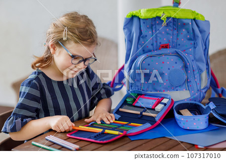 Happy smiling girl preparing for school her backpack. First day of school. Back to school concept. Little child collecting different supplies like pens and books in a bag or satchel. 107317010