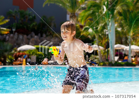Happy little kid boy jumping in the pool and having fun on family vacations in a hotel resort. Healthy child playing in water with a water gun. Laughing running child splashing Happy little kid boy jumping in the pool and having fun on family vacations in a hotel resort. Healthy child playing in water with a water gun. Laughing running child splashing 107317054
