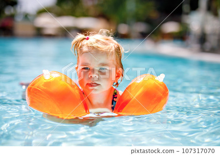 Little toddler girl with protective swimmies playing in outdoor swimming pool by sunset. Baby Child learning to swim in outdoor pool, splashing with water, laughing and having fun. Family vacations. 107317070