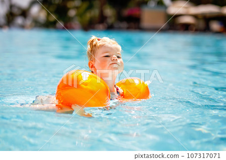 Little toddler girl with protective swimmies playing in outdoor swimming pool by sunset. Baby Child learning to swim in outdoor pool, splashing with water, laughing and having fun. Family vacations. 107317071
