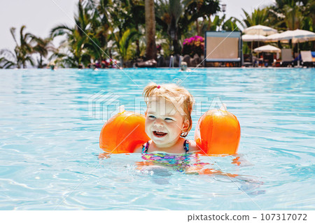 Little toddler girl with protective swimmies playing in outdoor swimming pool by sunset. Baby Child learning to swim in outdoor pool, splashing with water, laughing and having fun. Family vacations. 107317072