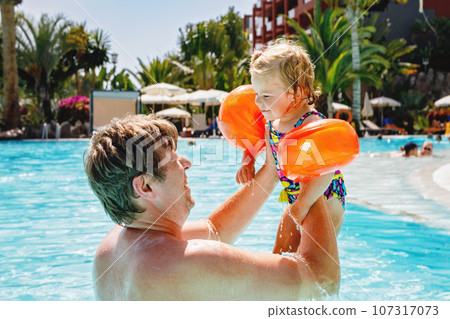 Cute happy little toddler girl and father in the pool and having fun on family vacations in a hotel resort. Healthy child and man playing in water. Baby daughter in colorful fashion swimsuit. 107317073