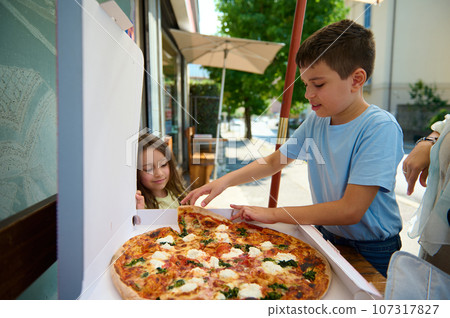 Adorable children, a teenage boy and little kid girl snacking outdoors, eating a delicious freshly baked Italian Margherita pizza with a crispy crust in the street. Italian lifestyle, cuisine, culture 107317827
