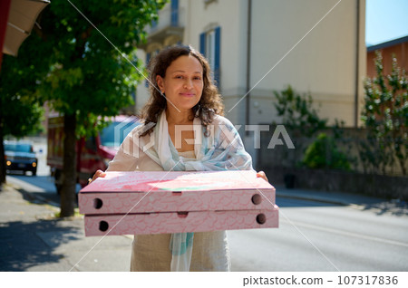 Beautiful curly haired multi-ethnic young woman in casual clothes, carrying two big disposable cardboard boxes with takeaway freshly baked Neapolitan pizza, smiles looking at camera. Italian lifestyle Beautiful curly haired multi-ethnic young woman in casual clothes, carrying two big disposable cardboard boxes with takeaway freshly baked Neapolitan pizza, smiles looking at camera. Italian lifestyle 107317836