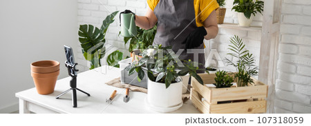 Relaxing home gardening banner. Smiling middle aged woman in black gloves with potted plant records gardening video blog in modern house - blogging and florist vlog influencer 107318059