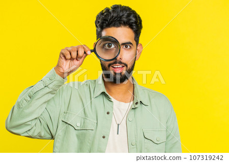 Indian young man holds magnifying glass near face looking at camera with big zoomed eye, analyzing 107319242
