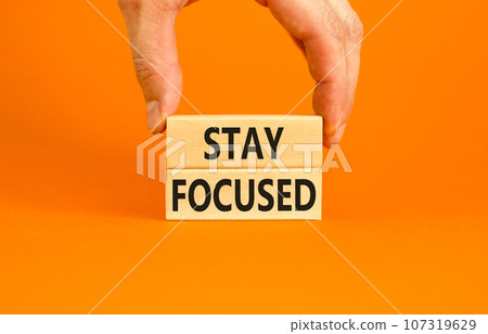 Stay focused symbol. Concept words Stay focused on wooden blocks on a beautiful orange background. Businessman hand. Business, support, motivation, psychological and stay focused concept. Copy space. Stay focused symbol. Concept words Stay focused on wooden blocks on a beautiful orange background. Businessman hand. Business, support, motivation, psychological and stay focused concept. Copy space. 107319629