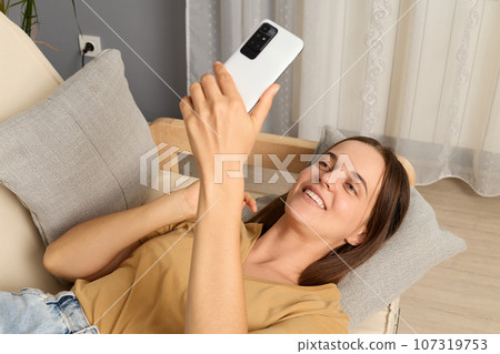 Digital leisure time. Web addiction. Social network chat. Online connection. Pretty cheerful positive brown haired woman wearing beige T-shirt lying on sofa holding smartphone in living room. Digital leisure time. Web addiction. Social network chat. Online connection. Pretty cheerful positive brown haired woman wearing beige T-shirt lying on sofa holding smartphone in living room. 107319753