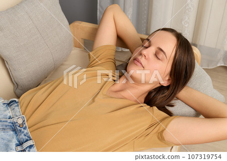 Relaxed young woman lying on couch holding hands behind head at home in modern living room napping keeps eyes close trying to have rest after hard working day. Relaxed young woman lying on couch holding hands behind head at home in modern living room napping keeps eyes close trying to have rest after hard working day. 107319754