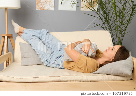 Depressed young adult Caucasian brown haired lying on sofa embracing pillow thinking about her problems and troubles felling loneliness looking away with pensive sad look. Depressed young adult Caucasian brown haired lying on sofa embracing pillow thinking about her problems and troubles felling loneliness looking away with pensive sad look. 107319755