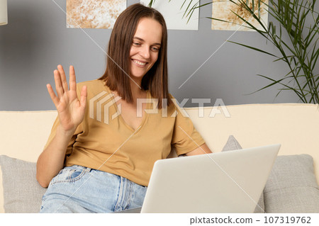Smiling cheerful brown haired woman wearing beige T-shirt and jeans sitting on couch and working on portable computer in home interior having video call looking smiling at screen waving hand. 107319762
