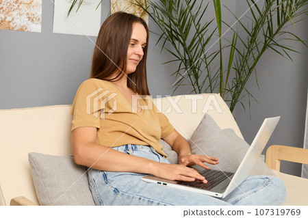 Remote occupation. Corporate networking. Employee productivity. Pleased pretty brown haired woman wearing beige T-shirt and jeans sitting on couch and working on portable computer in home interior Remote occupation. Corporate networking. Employee productivity. Pleased pretty brown haired woman wearing beige T-shirt and jeans sitting on couch and working on portable computer in home interior 107319769