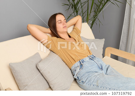 young sitting on sofa hands behind head at home in modern living room, enjoying a comfortable nap at home, keeps eyes closed, lost in a dream, taking moment to relax. young sitting on sofa hands behind head at home in modern living room, enjoying a comfortable nap at home, keeps eyes closed, lost in a dream, taking moment to relax. 107319792