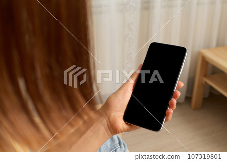 Mobile connection. Digital mockup. Virtual meeting. Unrecognizable brown haired woman holding smartphone with blank screen in hands light room interior. 107319801