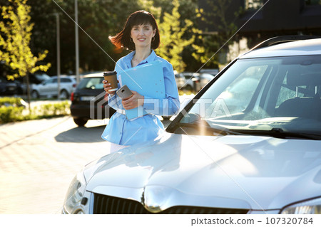 Positive confident businesswoman in elegant blue shirt holds folder with documents, cellphone and paper cup of hot drink looks at camera while standing front of her car by the modern office building 107320784
