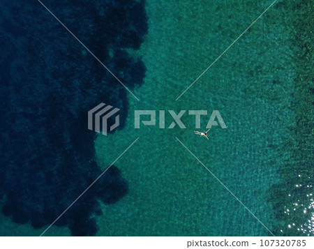 Above view of woman swimming at sea. Aerial top view of young girl in white swimsuit floating on water surface in crystal clear turquoise sea. Vacation at Paradise. Ocean relax, travel and vacation Above view of woman swimming at sea. Aerial top view of young girl in white swimsuit floating on water surface in crystal clear turquoise sea. Vacation at Paradise. Ocean relax, travel and vacation 107320785