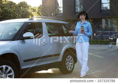 Elegant businesswoman in blue shirt, white pants holds paper cup of hot drink and smartphone while opening car by the modern office building. Female worker backs to home from work. Elegant businesswoman in blue shirt, white pants holds paper cup of hot drink and smartphone while opening car by the modern office building. Female worker backs to home from work. 107320786