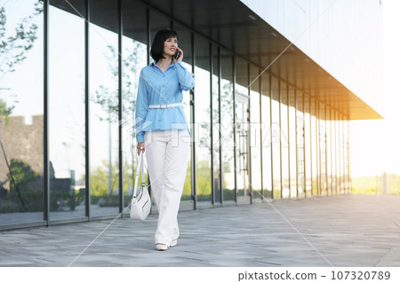 Successful elegant businesswoman entrepreneur or CEO in blue shirt and white pants smiles and talks on smartphone with partners or clients while walks by the office building outdoor. Business concept 107320789