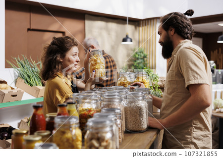 Farmer showcasing her products in zero waste marketplace location, selling chemicals free food to interested buyer. Local trader selling organic farm grown bulk products during event to client 107321855