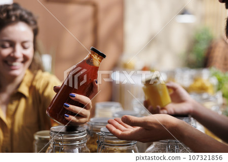 Woman shops in zero waste grocery store, looks at bulk products in reusable packaging while being assisted by happy storekeeper. Trader in eco friendly shop recommending food item to client, close up 107321856