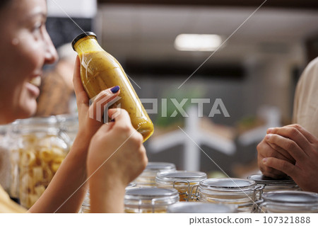 Vendor showcasing her products in zero waste marketplace venue, selling homemade fruit juice to interested customer, close up. Local farmer trading healthy food items during event, blurry background Vendor showcasing her products in zero waste marketplace venue, selling homemade fruit juice to interested customer, close up. Local farmer trading healthy food items during event, blurry background 107321888